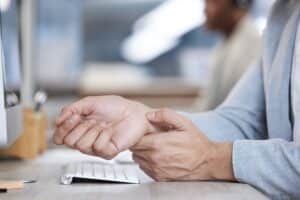 Businessman experiencing wrist pain while working on a computer, highlighting carpal tunnel syndrome and the need for physical therapy or stress relief for repetitive strain injuries.