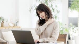 Woman experiencing stiff shoulders while working on a computer, considering whiplash physical therapy for relief.
