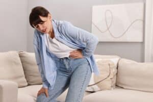 Upset woman at home holding her hip in discomfort, showing signs of hip joint pain and limited mobility.